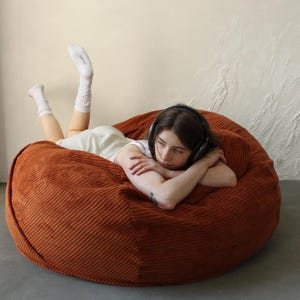 May include: A large, round, rust-colored corduroy beanbag chair. The beanbag is the focal point, with a person wearing headphones and white socks lying on it. The background is a neutral beige and white.