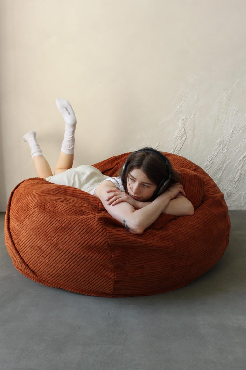 May include: A large, round, rust-colored corduroy beanbag chair. The beanbag is the focal point, with a person wearing headphones and white socks lying on it. The background is a neutral beige and white.