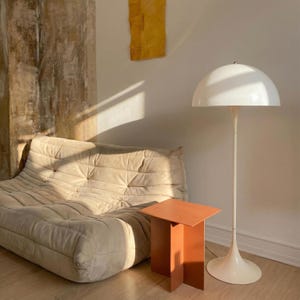 May include: A cream-colored floor lamp with a white, dome-shaped shade stands next to a small, orange side table. A beige, tufted sofa is on the left. A piece of yellow art hangs on the wall.