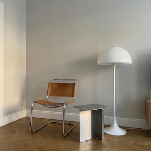 May include: A modern interior scene featuring a brown leather chair with a chrome frame, a small brushed metal side table, and a white floor lamp with a dome-shaped shade. The room has light gray walls and a wooden parquet floor.