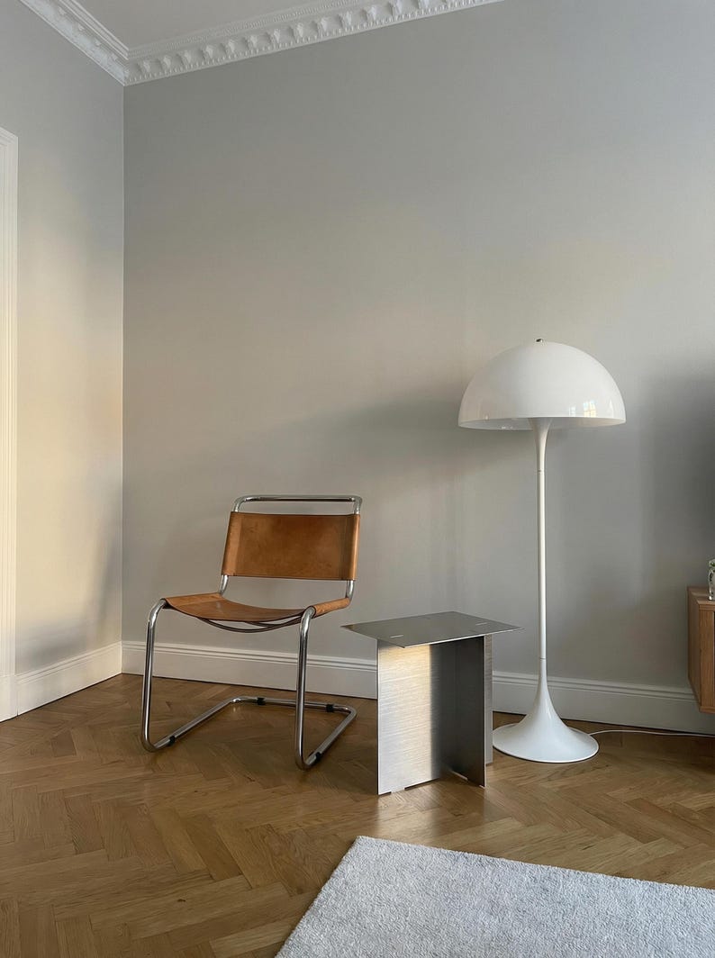 May include: A modern interior scene featuring a brown leather chair with a chrome frame, a small brushed metal side table, and a white floor lamp with a dome-shaped shade. The room has light gray walls and a wooden parquet floor.