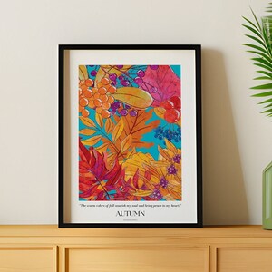 Op de afbeelding: Een ingelijst drukwerk met een kleurrijke illustratie van herfstbladeren en bessen op een blauwe achtergrond. De tekst "The warm colors of fall surround me, and still bring peace to my heart. AUTUMN" is onder de illustratie gedrukt.