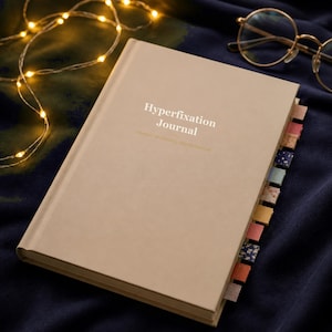 May include: A beige hardcover journal with the text Hyperfixation Journal printed in white on the front cover. The journal features multiple colorful patterned page markers extending from the side and is accompanied by gold-rimmed round glasses.