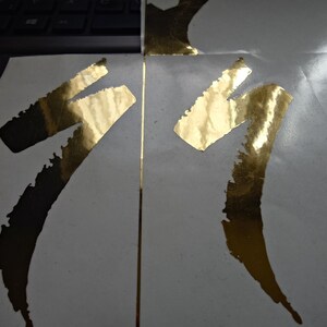 May include: Close-up of two sections of a white surface with gold foil accents. The gold foil is applied in a stylized, abstract shape. The image is well-lit, highlighting the texture and sheen of the gold foil against the white background.