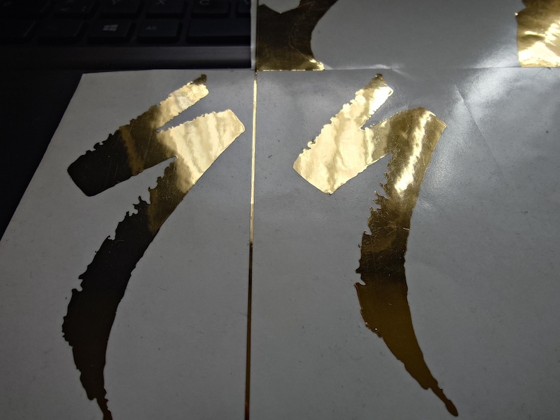 May include: Close-up of two sections of a white surface with gold foil accents. The gold foil is applied in a stylized, abstract shape. The image is well-lit, highlighting the texture and sheen of the gold foil against the white background.