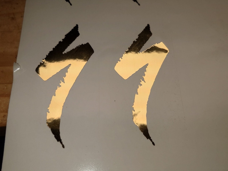 May include: Gold metallic decals in a stylized, abstract design. The decals are in the shape of a number seven, with a textured, torn edge. The decals are on a white background.