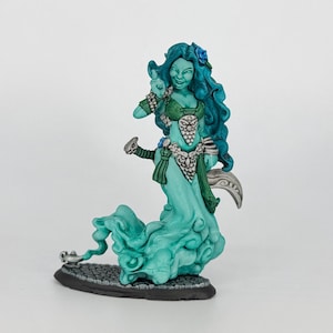 May include: A teal-colored fantasy figurine of a female figure with long, flowing teal hair and a matching dress. The figure has a green top and silver accents. She is holding a curved blade and is standing on a black base.