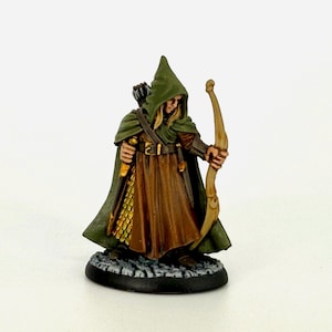 May include: Miniature figure of an archer in a green hooded cloak and brown tunic, holding a bow. The figure has blonde hair and is standing on a textured base. The bow is drawn, ready to fire an arrow. The figure is painted with detailed features.