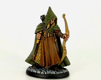 May include: Miniature figure of an archer in a green hooded cloak and brown tunic, holding a bow. The figure has blonde hair and is standing on a textured base. The bow is drawn, ready to fire an arrow. The figure is painted with detailed features.