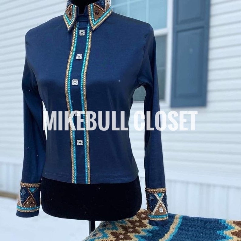 May include: A navy blue long-sleeved shirt with decorative embellishments. The shirt features a high collar and cuffs with brown, gold, and turquoise accents. The front has a vertical turquoise and gold trim with square silver buttons. The text "MIKEBULL CLOSET" is across the front.