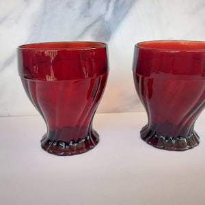 Könnte beinhalten: Zwei rote Glasbecher mit geriffeltem Design. Die Gläser haben einen breiten Boden und eine abgerundete Oberseite.