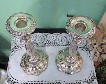 May include: Two silver-colored candle holders with ornate floral designs. The candle holders have a round base and a detailed stem leading to a circular top. The tops have a recessed area for holding candles.