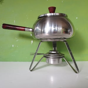 Può includere: Una fonduta d'argento con un manico in legno e un supporto con un bruciatore. La pentola ha un coperchio con un pomello in legno.