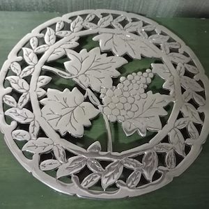 May include: Circular silver-colored trivet with a grape and leaf design. The trivet features a central cluster of grapes surrounded by large leaves and a border of smaller leaves. The metal trivet is designed to protect surfaces from hot dishes.