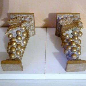 May include: Two gold-painted decorative wall brackets shaped like grapes. Each bracket features a cluster of grapes with leaves and a base.
