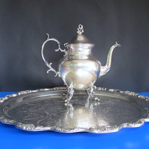 Puede incluir: Una tetera de plata con una tapa decorativa y un asa curva se encuentra sobre una bandeja de plata con un borde festoneado. La bandeja tiene un diseño floral.