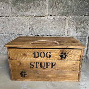 May include: A wooden storage box for dog items, with a lid and rope handle. The box is made of light brown wood and features the words "DOG STUFF" in black, along with paw print designs.