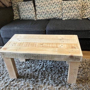 May include: A rustic wooden coffee table with a natural wood finish. The table has a rectangular top and four legs. The table is sitting on a light brown rug.