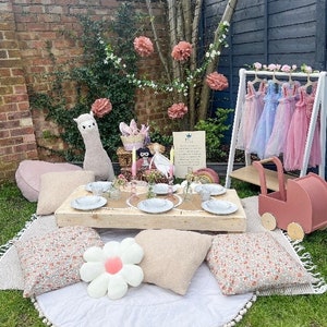 May include: A picnic set up with a wooden table, white plates, and a pink and white floral tablecloth. There are pink and white cushions, a white daisy cushion, and a pink toy pram. The background features a brick wall, a wooden fence, and a tree with pink paper flowers.
