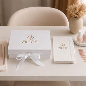 May include: A white rectangular gift box with a gold logo reading "aires" and a white ribbon bow, alongside a matching white notebook with the same gold logo, a white pen, two small bottles, and a fan of color swatches on a white surface.