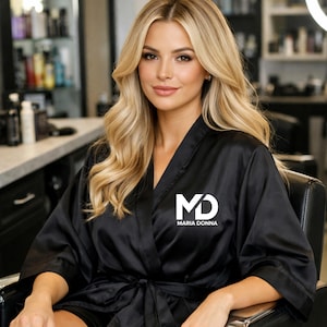 May include: Black satin robe with the white logo "MD MARIA DONNA". The robe is tied at the waist and has wide sleeves. The model has long blonde hair and is sitting in a black chair.