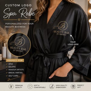 May include: A black satin spa robe featuring a custom gold embroidered logo on the chest that reads Beauty Bar by Bruna. The robe includes a matching tie belt, wide sleeves, and two front pockets. Text on the image includes Custom Logo Spa Robe.