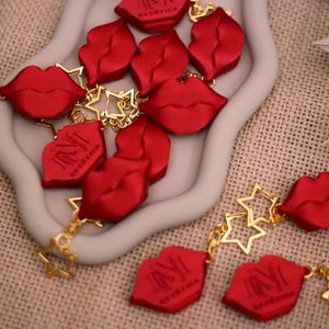 Puede incluir: Una colección de dijes con forma de labios rojos con acabado mate, algunos con el texto "INVIT ESTÉTICA" en negro. Los dijes están conectados por cadenas de metal dorado y pequeños eslabones en forma de estrella abierta.