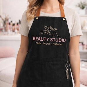 May include: Black apron with brown leather straps and gold rivets. The apron has a pocket and a key ring with tools. The words "BEAUTY STUDIO" are printed in pink, with "nails • brows • esthetics" below.