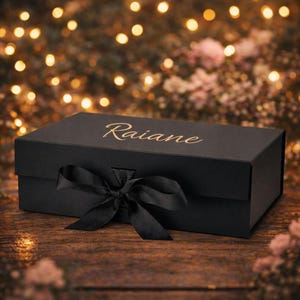 May include: A black gift box with a matching black ribbon tied in a bow. The box has the name "Raiane" in gold script on the lid. The box is on a wooden surface with a blurred background of lights and flowers.