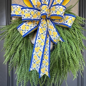 May include: A decorative wreath featuring a lush green base and a large bow. The bow is made of blue and yellow ribbon with a floral pattern. The wreath is likely used for seasonal or holiday decor.