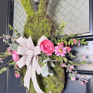 Pu&ograve; includere: Una ghirlanda a forma di coniglio ricoperta di muschio, con un fiocco a righe e fiori rosa. La ghirlanda &egrave; decorata con verde artificiale e fiori rosa, creando una decorazione primaverile o pasquale. Appesa a una porta scura.