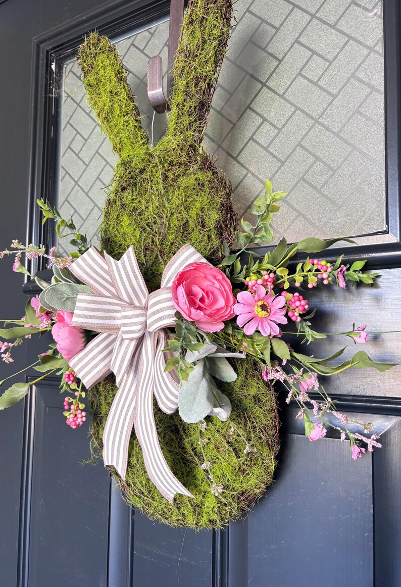 Pu&ograve; includere: Una ghirlanda a forma di coniglio ricoperta di muschio, con un fiocco a righe e fiori rosa. La ghirlanda &egrave; decorata con verde artificiale e fiori rosa, creando una decorazione primaverile o pasquale. Appesa a una porta scura.