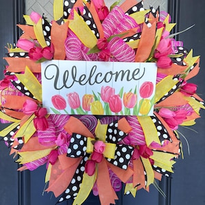 May include: A vibrant wreath with pink, orange, yellow, and black ribbons and faux tulips. A white sign in the center reads "Welcome" above a row of tulip illustrations. The wreath is designed for a front door.
