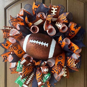 May include: A decorative wreath featuring a central brown glittery football surrounded by mesh ribbons in navy blue and orange. The ribbons display patterns of tiger stripes, paw prints, and football field markings with small football accents.