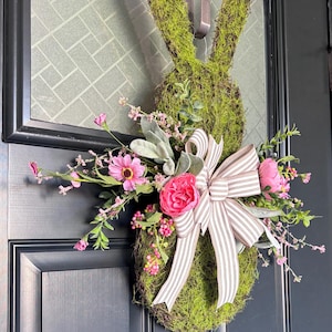 Pu&ograve; includere: Una ghirlanda a forma di coniglio ricoperta di muschio con un fiocco decorativo e fiori rosa. La ghirlanda &egrave; appesa a una porta nera, creando una decorazione festosa pasquale o primaverile.