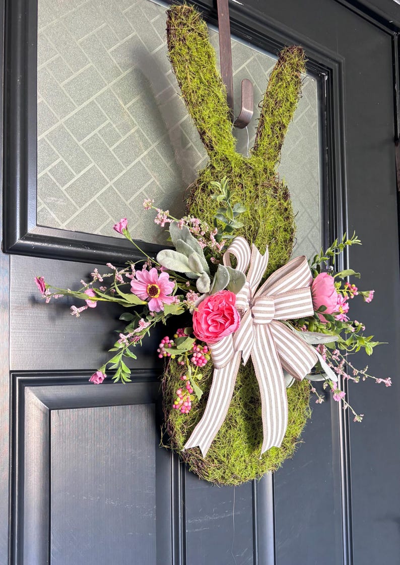 Pu&ograve; includere: Una ghirlanda a forma di coniglio ricoperta di muschio con un fiocco decorativo e fiori rosa. La ghirlanda &egrave; appesa a una porta nera, creando una decorazione festosa pasquale o primaverile.