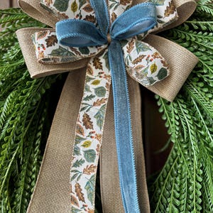 May include: A decorative bow featuring layered ribbons of tan burlap, a white ribbon with a colorful autumn leaf and berry pattern, and a central blue velvet ribbon, all secured atop a wreath of dense, textured green fern-like foliage.