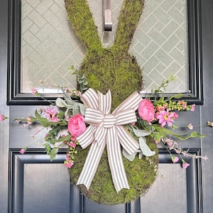 Pu&ograve; includere: Una ghirlanda a forma di coniglio ricoperta di muschio, con un fiocco a righe e fiori rosa. La ghirlanda &egrave; appesa a una porta nera. Il coniglio ha lunghe orecchie ed &egrave; decorato con fiori rosa e verde.