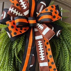 May include: A decorative bow featuring a mix of ribbons with black and orange polka dots, solid black and orange stripes, and brown football patterns with white laces. The bow is layered with multiple loops and long trailing ribbon tails.