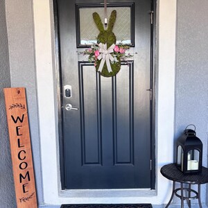 Pu&ograve; includere: Una ghirlanda a forma di coniglio verde scuro con fiori rosa e un fiocco bianco &egrave; appesa a una porta d'ingresso nera. Sono visibili anche un cartello di benvenuto in legno e una lanterna nera su un tavolino.