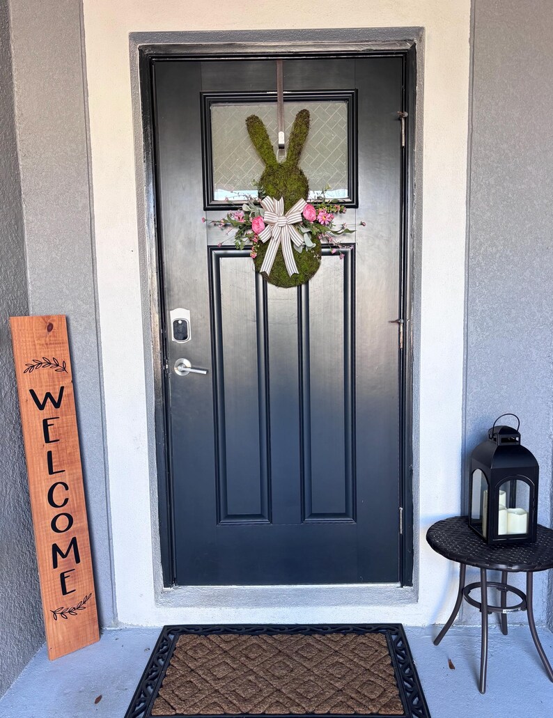 Pu&ograve; includere: Una ghirlanda a forma di coniglio verde scuro con fiori rosa e un fiocco bianco &egrave; appesa a una porta d'ingresso nera. Sono visibili anche un cartello di benvenuto in legno e una lanterna nera su un tavolino.