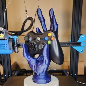 May include: A 3D-printed, blue hand-shaped game controller holder. The holder is designed to cradle a black Xbox controller. The base is white and circular, providing stability. The design is intricate and detailed, showcasing the texture of the 3D printing process.