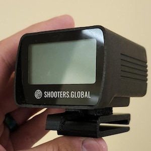 May include: A black electronic device with a digital display and the text "SHOOTERS.GLOBAL" on the front. The device has a clip on the bottom.