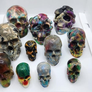 May include: A collection of eleven decorative skull sculptures in various sizes and colors. The skulls feature a glossy finish and are painted with a variety of colors including red, blue, green, and yellow. The skulls are arranged on a white surface.