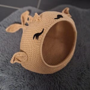 May include: A beige crocheted cat bed shaped like a cat's head with black embroidered eyes and ears.