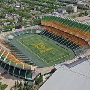 May include: Aerial view of a large outdoor stadium with green and gold seating. The field is green with a gold logo at the center. The stadium is surrounded by buildings and trees.