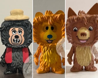 May include: Three hand-painted figurines of cartoon animals. The first is a black monkey wearing a hat and scarf. The second is an orange lion. The third is a brown fox with pointed ears. All have smiling faces.
