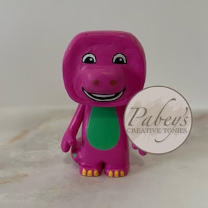 Könnte beinhalten: Eine lila Dinosaurierfigur mit grünem Bauch und gelben Füßen. Die Figur hat ein lächelndes Gesicht und hält ein weißes rundes Schild mit dem Text "Pabey's Creative Tonies".