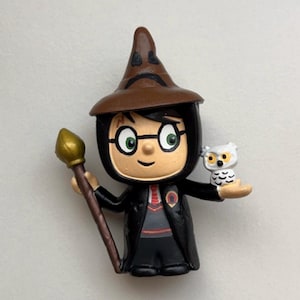 May include: A small plastic figurine of Harry Potter wearing a brown pointed hat, black robes, and glasses. He is holding a brown wand and a white owl.