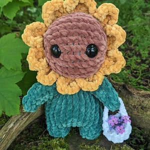 May include: A crocheted sunflower plush toy with a green body and a brown face. The toy is holding a white crocheted basket with purple flowers.
